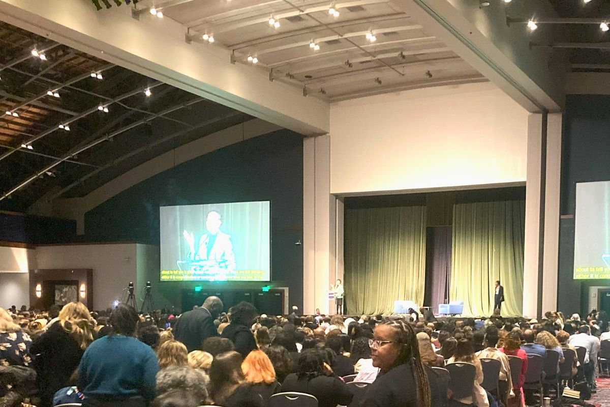 Kimberle Crenshaw stands on stage delivering the opening plenary. Attendees fill the chairs in a large room, and some attendees sit on the floor.